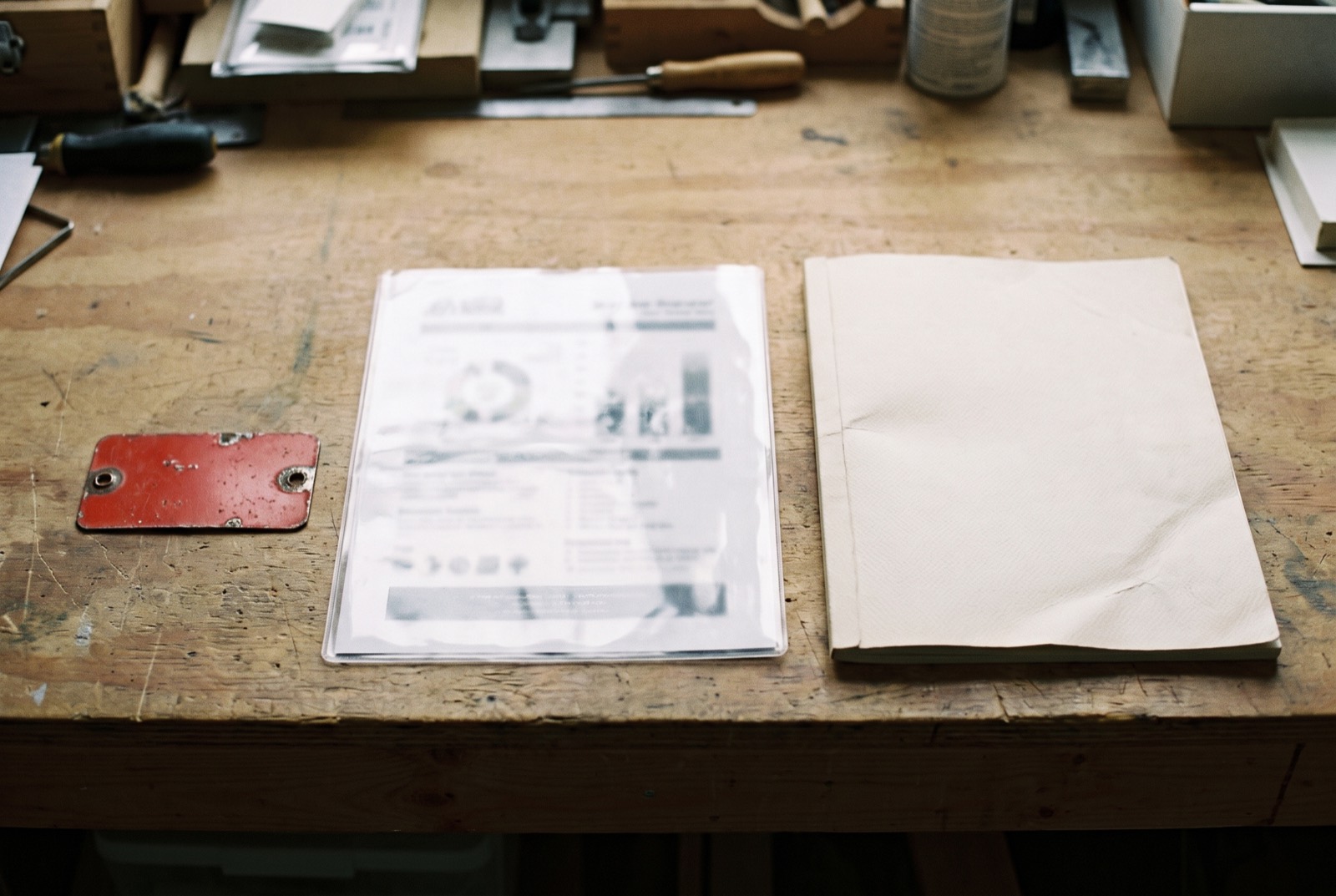 A bound report, a printed sheet in a plastic sleeve and a small red metal tag on a workbench